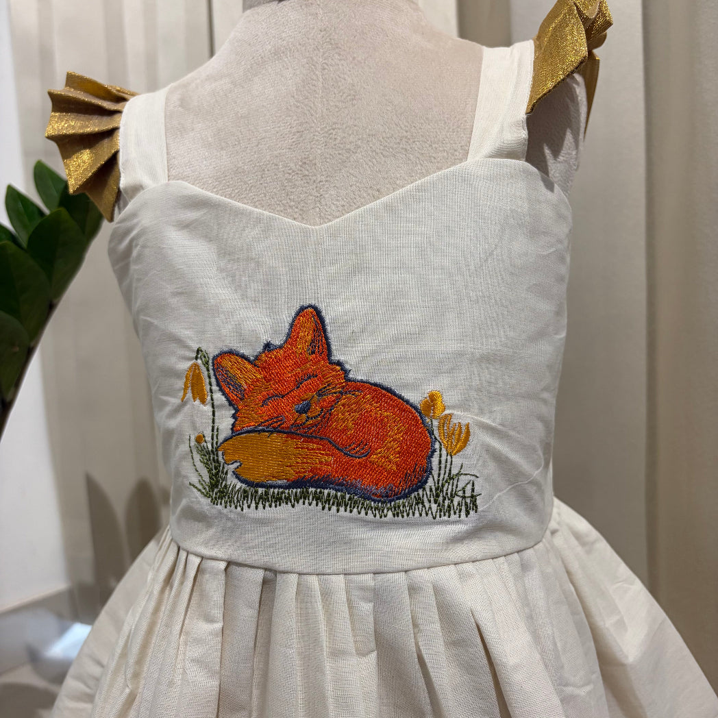 An off-white sleeveless dress displayed on a mannequin, featuring a colorful embroidered sleeping fox in orange and yellow with green grass on the bodice. The dress has gold ribbon shoulder straps and a gathered skirt.