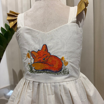 An off-white sleeveless dress displayed on a mannequin, featuring a colorful embroidered sleeping fox in orange and yellow with green grass on the bodice. The dress has gold ribbon shoulder straps and a gathered skirt.
