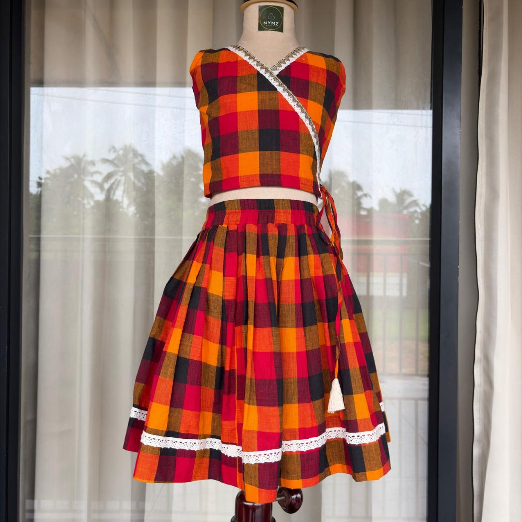 A sleeveless two-piece plaid set on a mannequin, featuring a wrap-style crop top and a matching pleated skirt in red, orange, yellow, and black, with white lace trim details.