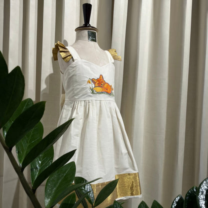 A white sleeveless dress displayed on a mannequin torso indoors, with gold shoulder bows, a small orange and yellow embroidered motif at the center, and a gold hem detail. The dress is photographed in front of beige curtains with large green plant leaves partially obscuring the foreground.