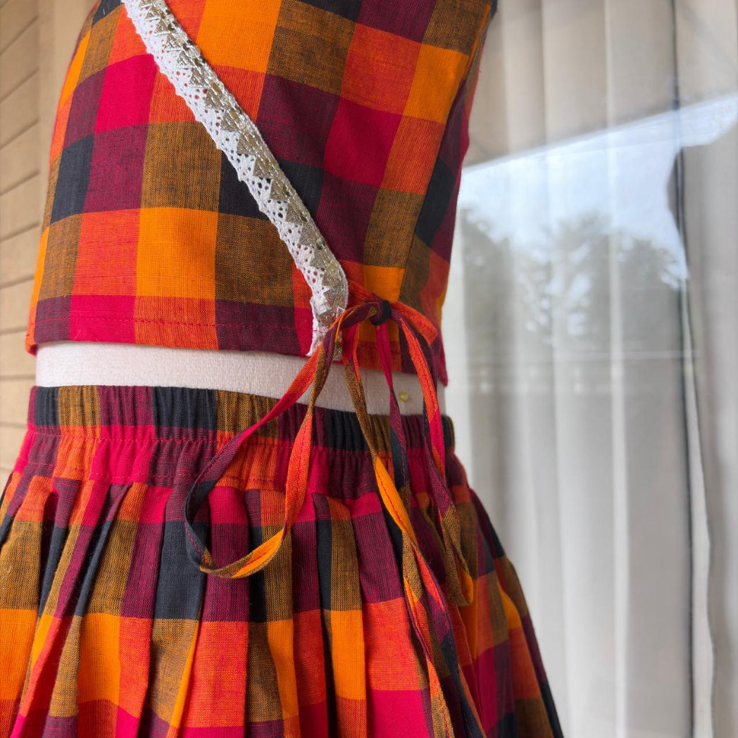 A close-up of a plaid dress or skirt in warm autumn colors including orange, red, brown, and black, with a white lace strap detail and elastic waist tied with ribbon.
