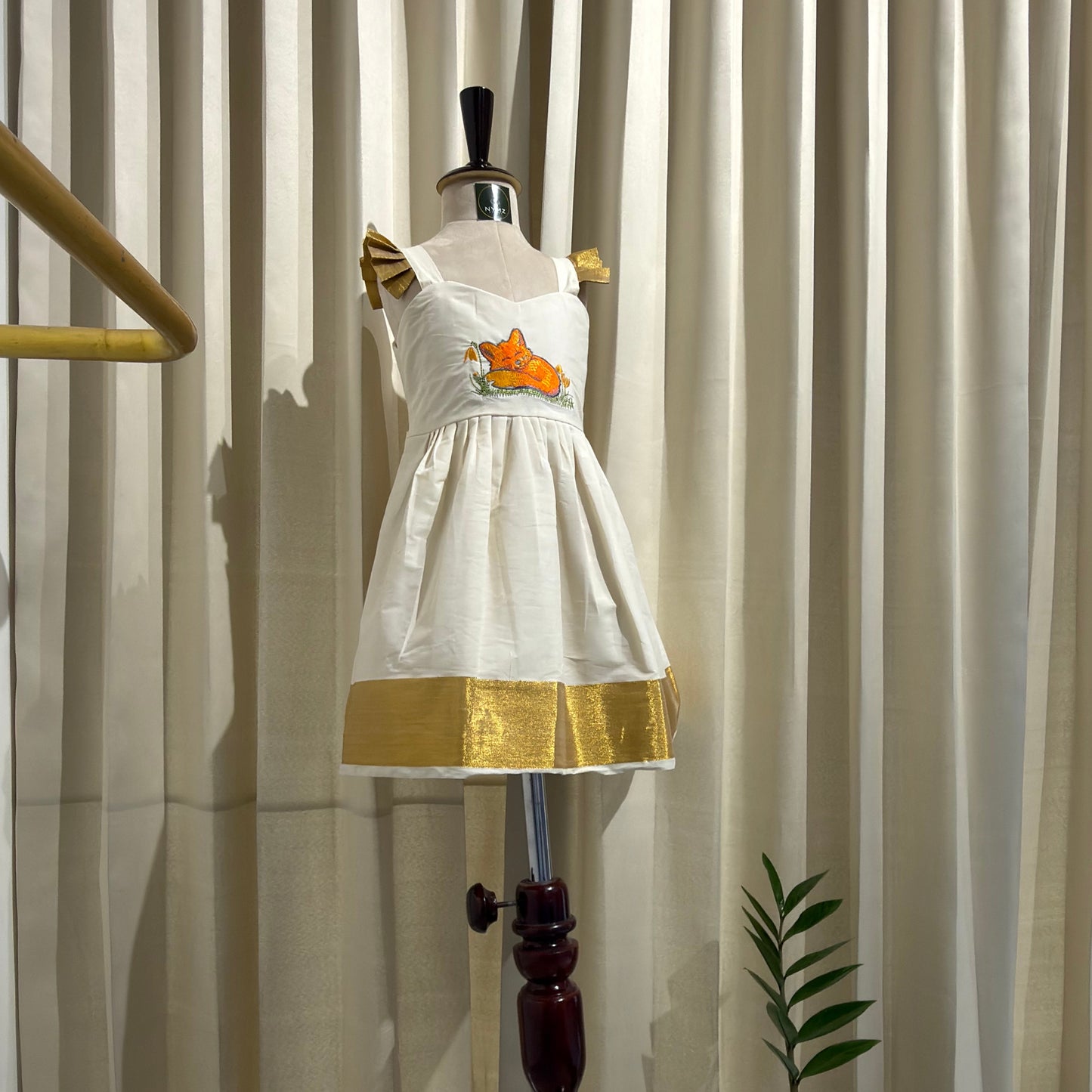 A white sleeveless dress or tunic with a gold band and small orange fox graphic on the chest, displayed on a dress form mannequin against a beige pleated curtain backdrop.